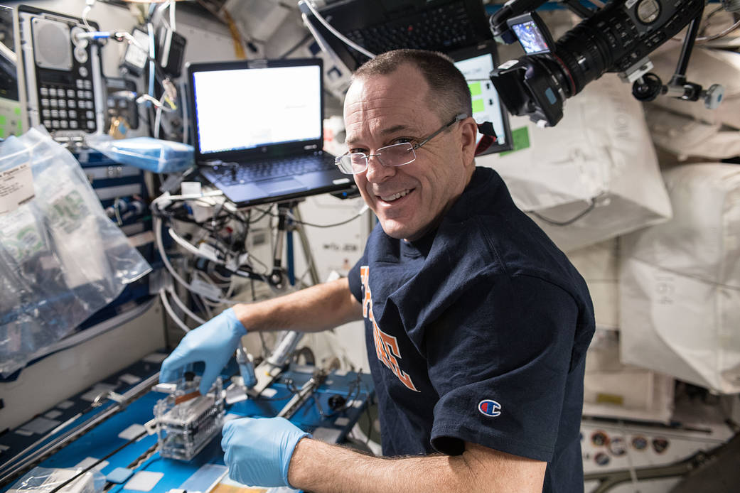 Arnold, facing the camera, wears a blue shirt, glasses, and light blue gloves. His right hand holds the miniPCR on the work bench. A laptop and video camera are visible behind him.