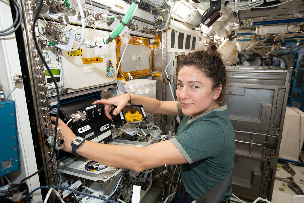NASA astronaut Jessica Meir is positioned in front of an open a compartment on a wall of the space station. Inside is a black box-shaped device, about the size of a large watermelon. Meir’s body is facing towards the black device as she adjusts it. Her head is turned to the camera with a subtle smile.