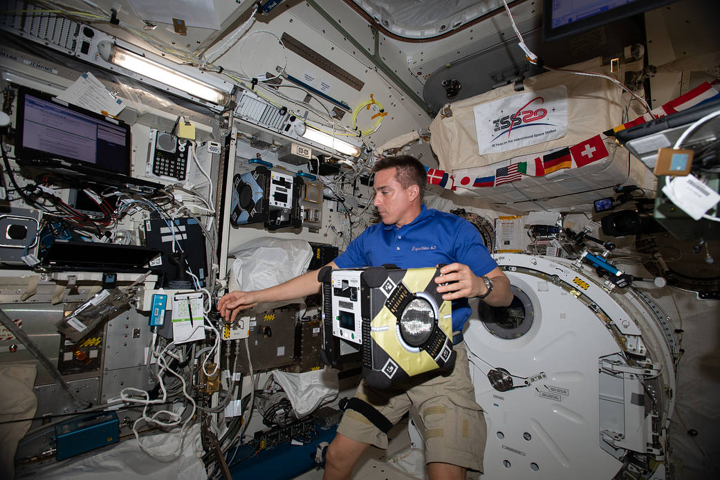 Cassidy, wearing a blue polo shirt and khaki shorts, holds the yellow Astrobee in his left hand as he reaches for a clipboard with his right hand. Also visible is the blue Astrobee in the docking station to his right, a round white hatch behind him, and a string of small flags just above his head.