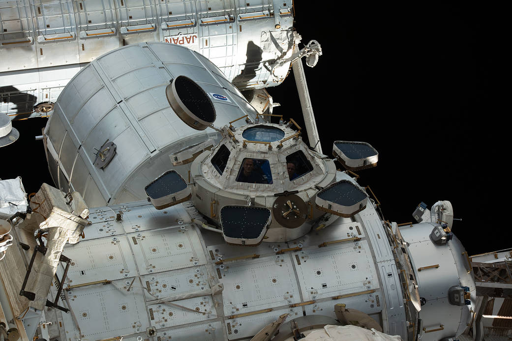 Expedition 66 crew members peer out from windows inside the cupola