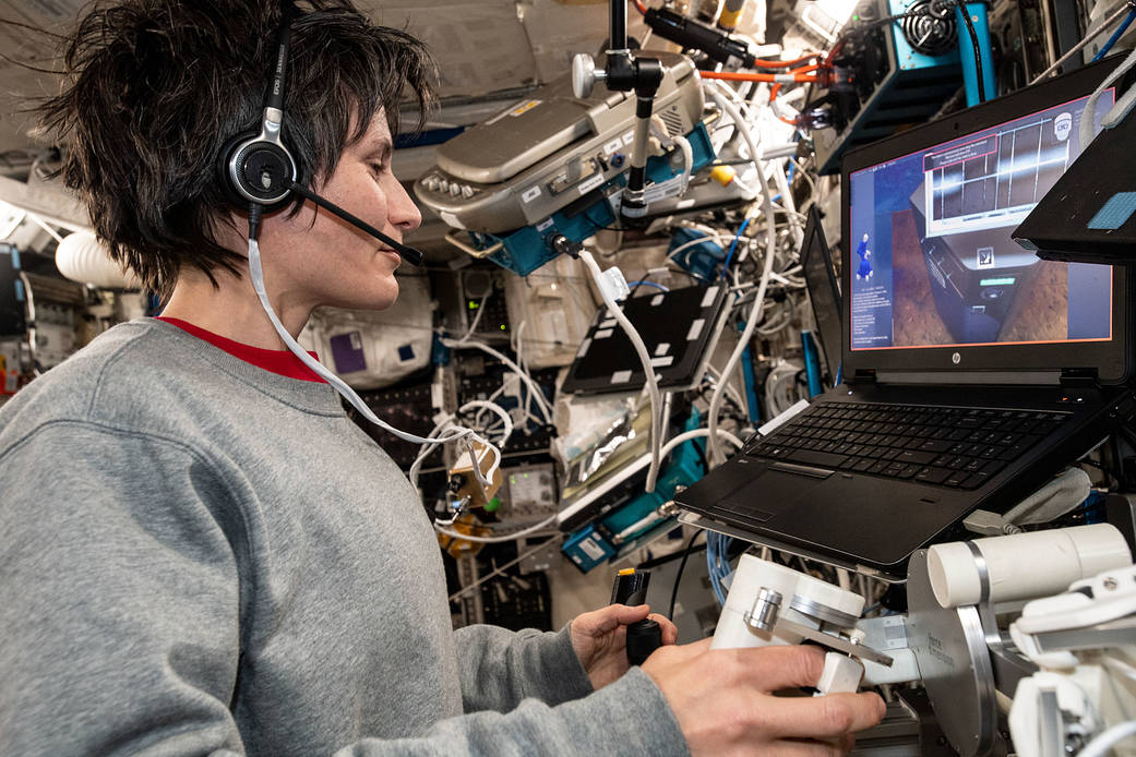 Cristoforetti, wearing a gray sweatshirt and a headset, looks at a computer screen in front of her as she works a joystick with her left hand and holds a white control arm with her right hand.