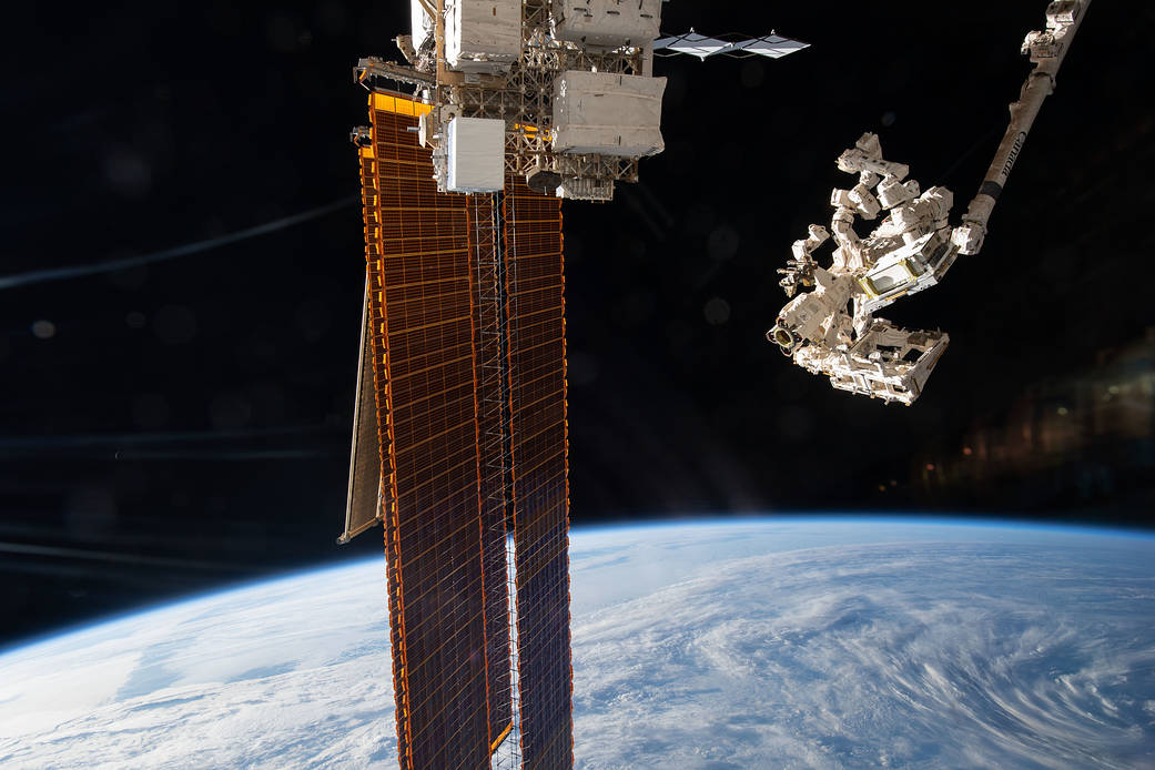 The Canadarm2 robotic arm maneuvers the Earth Surface Mineral Dust Source Investigation (EMIT) toward an ExPRESS Logistics Carrier-1 (top center) on the International Space Station's port side truss structure. The station's main solar arrays are also seen extending from the port truss segment.