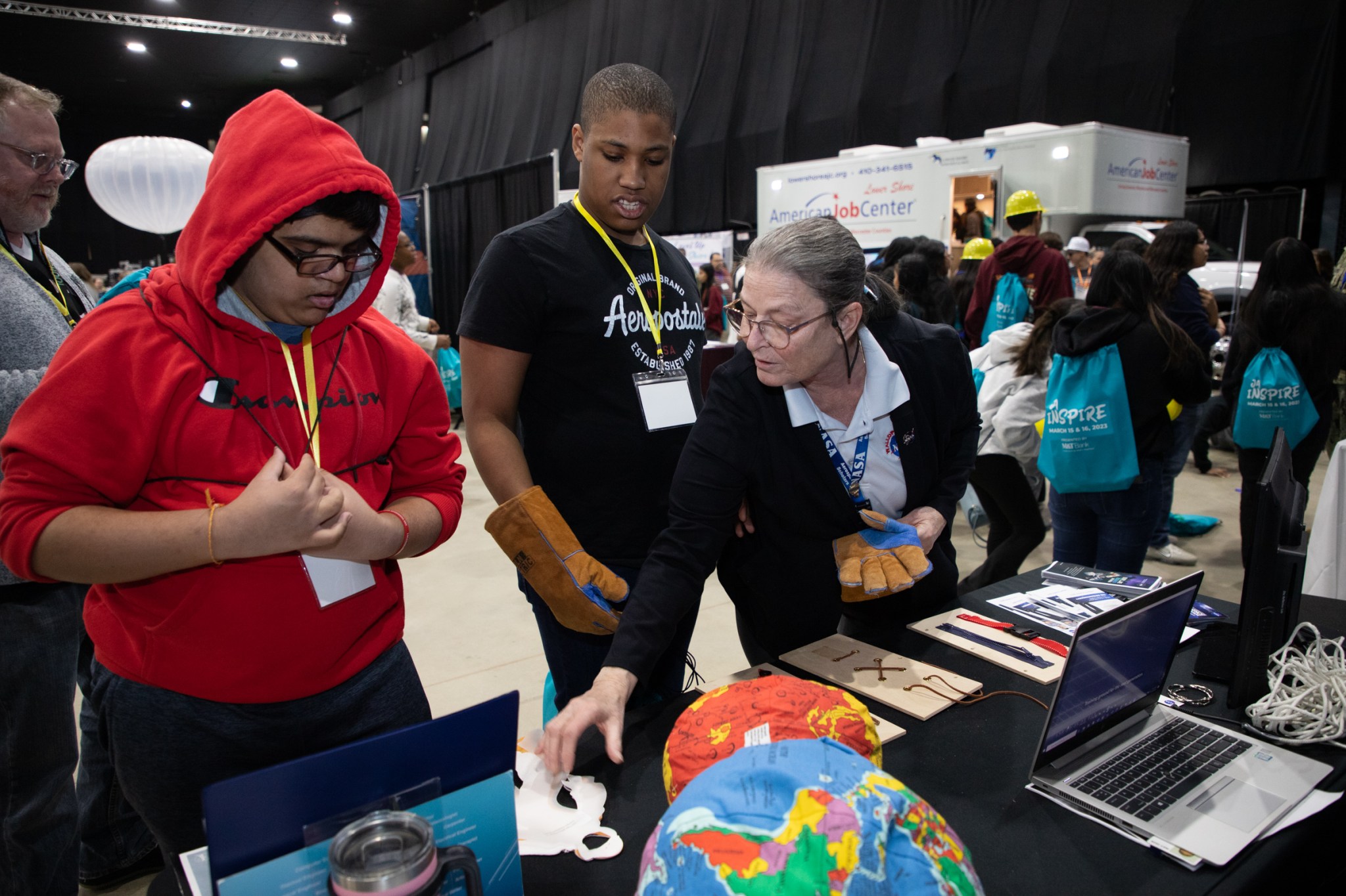 A woman points at educational materials set out on a table while two students look down at the table. Behind them are other booths at a convention center.