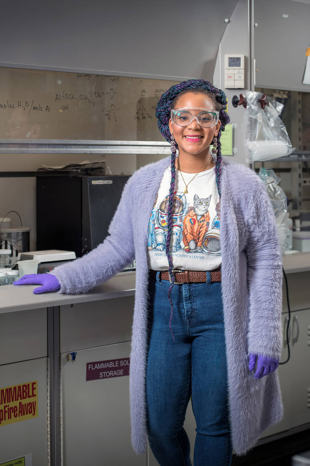 Young, black woman with safety glasses, dressed in t-shirt, jeans, and purple fuzzy sweater with purple gloves, faces camera while standing in laboratory