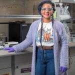 Young, black woman with safety glasses, dressed in t-shirt, jeans, and purple fuzzy sweater with purple gloves, faces camera while standing in laboratory