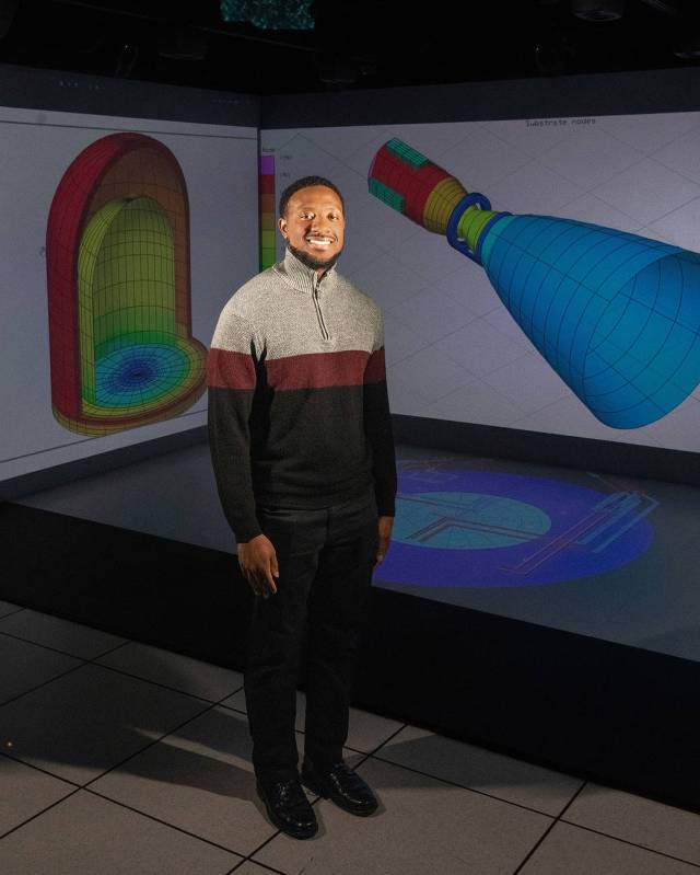 Man in sweater stands in front of 3D technical drawings while looking at the camera.