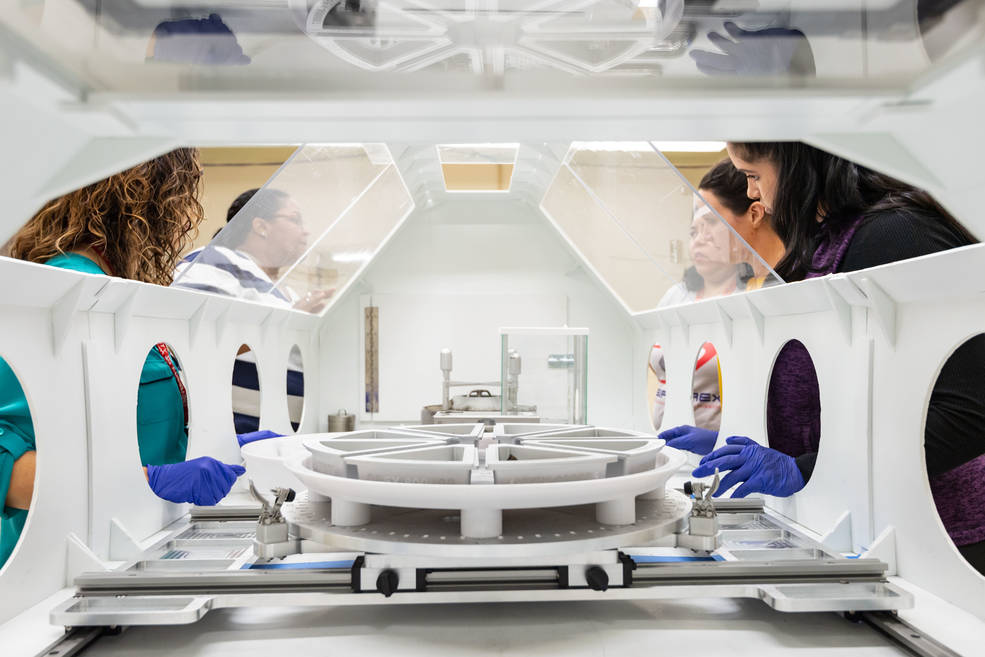 Interior view of a white glove box for use in analyzing samples. Technicians' faces are visible through glass panels on the box