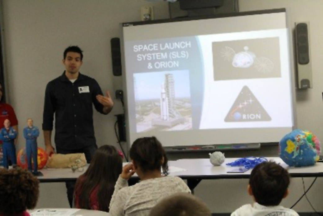 Johnson intern Kaylon Wheeler stands in front of a local classroom as he talks to students about NASA. Beside him, a screen show