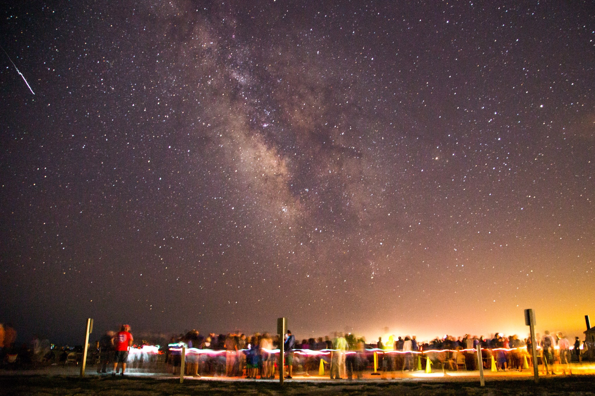 Hundreds of people stand in lines on the beach waiting their turn to look through telescopes at an astronomy event, above them the cloudy streaks of the milky way are surrounded by thousands of bright stars.