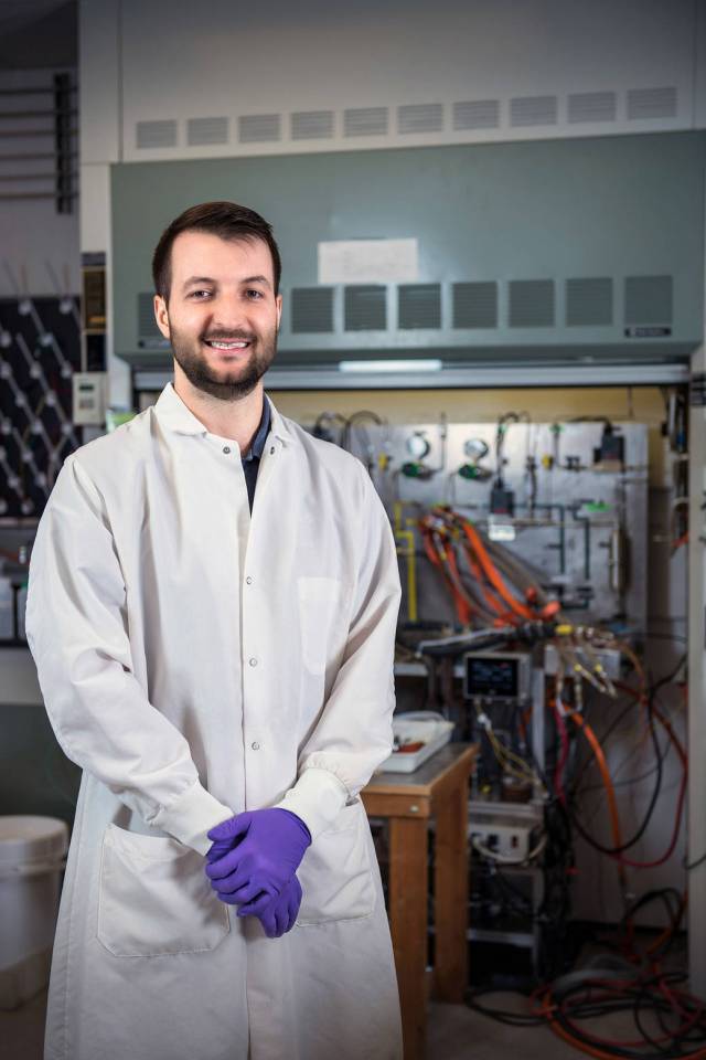 Young man in lab coat and purple protective gloves smiles at camera inside lab filled with electronics cables.