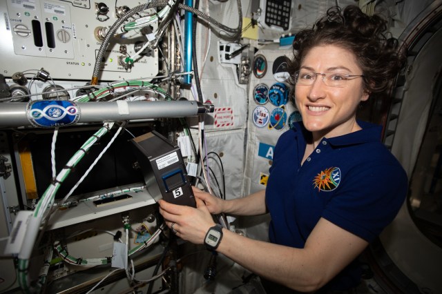 Female astronaut in black shirt with brown hair posing for a photo while working with experiment hardware aboard the space station.