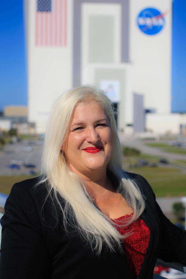 A woman wearing a black blazer, red blouse, and red lipstick smiles softly while looking at the camera. NASA's Vehicle Assembly Building stands behind her. 