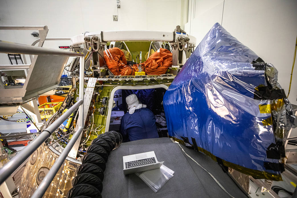 A technician with a protective covering over their head and body work inside the Orion capsule from Artemis I. A laptop is the foreground while deflated orange crew module uprighting system bags are atop the capsule.