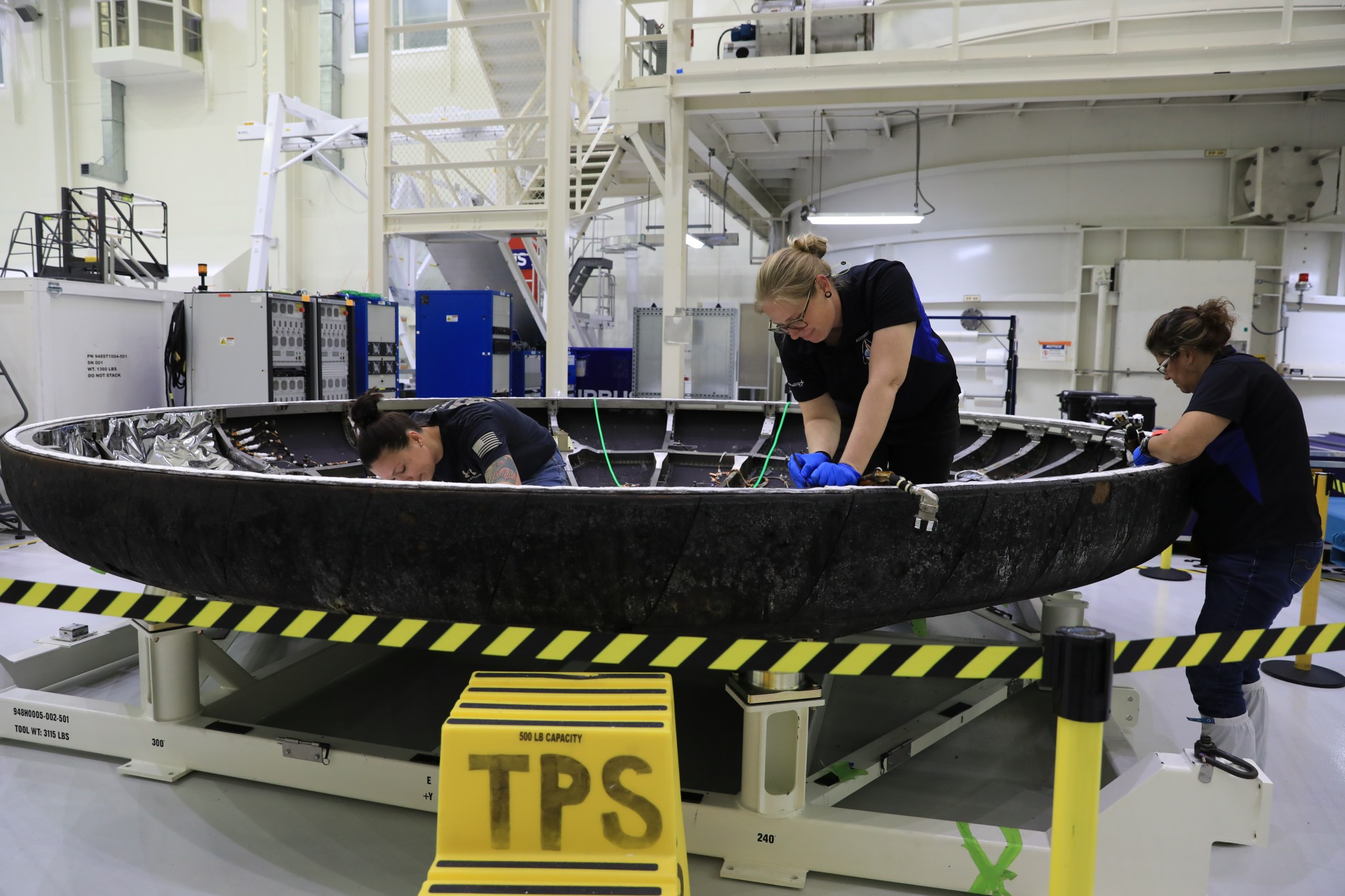 Technicians examine the black Orion heat shield, detached from the rest of the spacecraft and on a stand.