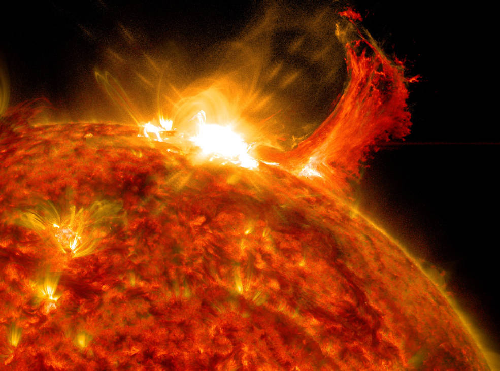 A close-up view shows part of the Sun in red, orange, and yellow. A bright yellow-white burst of light appears at the edge of the Sun in the top center. To the right of that, a red, column-shaped cloud of gas towers above the edge of the Sun.