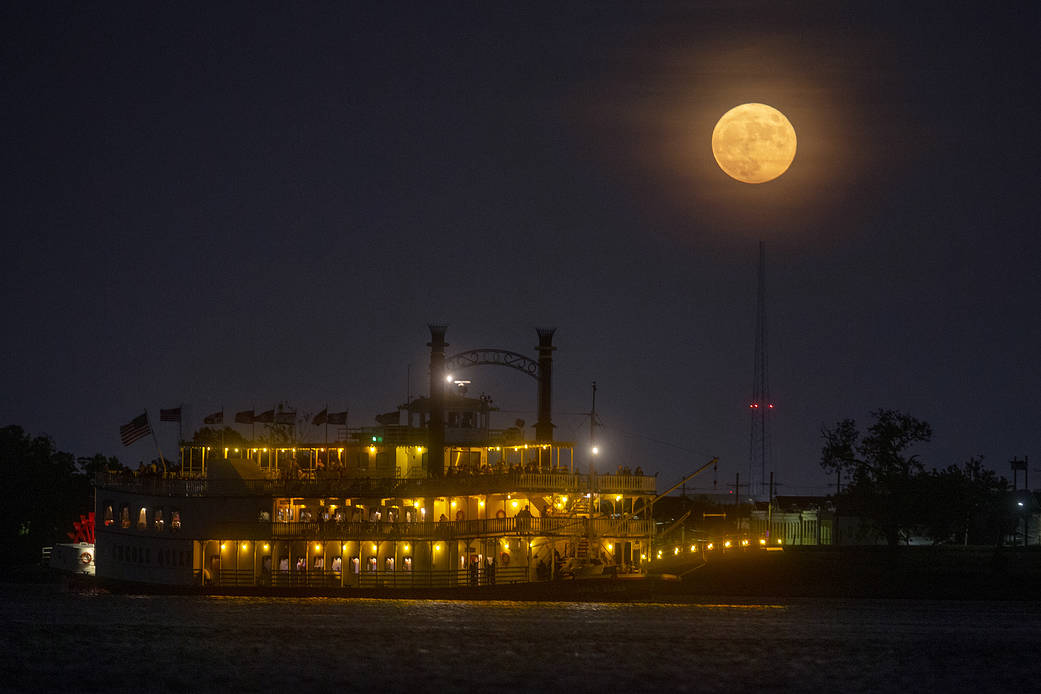 An orange-yellow full Moon is in the night sky above a lit-up riverboat.