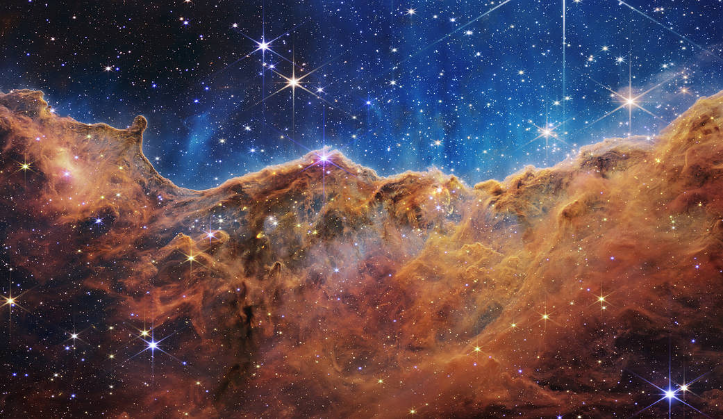 an undulating, translucent star-forming region in the Carina Nebula is shown in this Webb image, hued in ambers and blues; foreground stars with diffraction spikes can be seen, as can a speckling of background points of light through the cloudy nebula