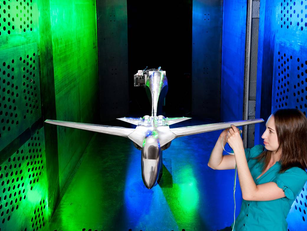 Small silver aircraft model in wind tunnel with researcher adjusting wing at right