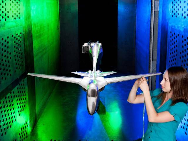 Small silver aircraft model in wind tunnel with researcher adjusting wing at right