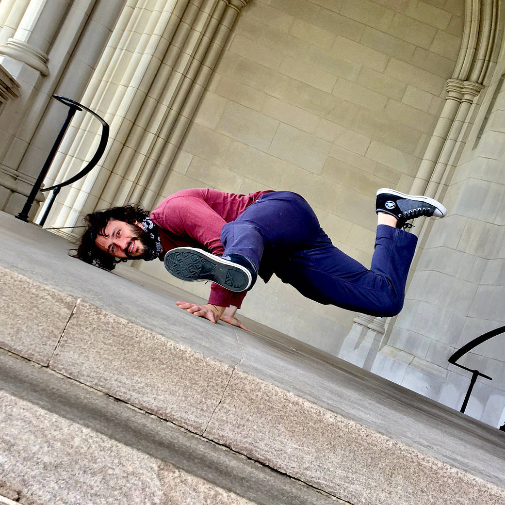 Dr. Mason Quick, a man with long brown hair, a beard, and mustache, wears a red long sleeve shirt, blue slacks, and black and grey converse. He is holding himself up on his hands, with his body horizontal to the ground and legs kicked out in a split.