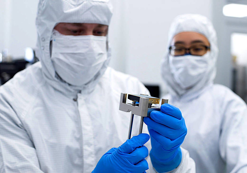 A man wearing a white suit, white face mask, and blue gloves holds one of Roman's detectors, which is about as wide as a Saltine cracker and half an inch thick. A woman (also wearing a white suit and face mask) watches and supervises.