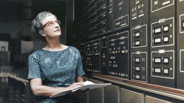 Portrait of astronomer Nancy Grace Roman holding papers and looking up at a board with information on it