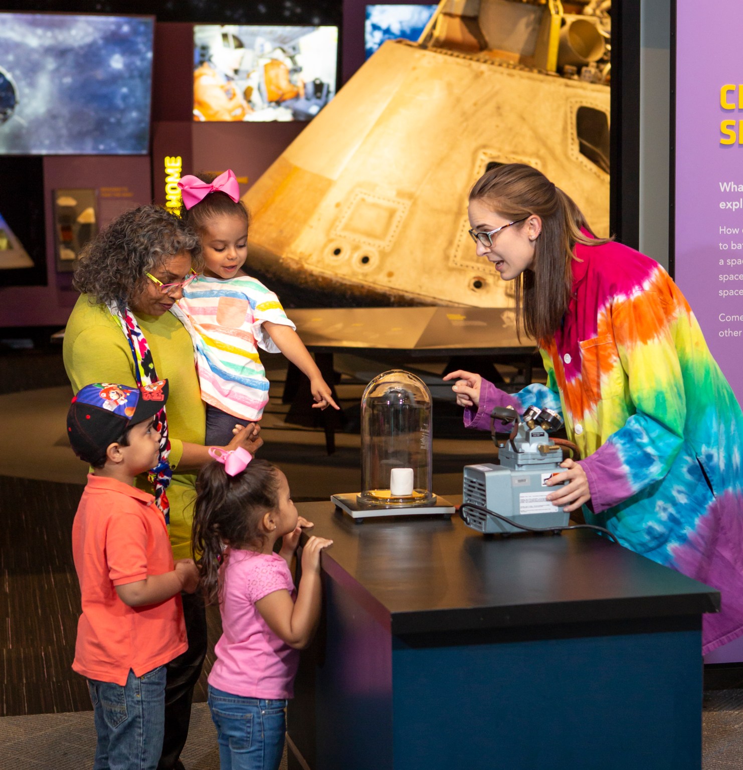 A woman wearing a colorful lab coat demonstrates a science project for a mother and children at the Great Lakes Science Center.