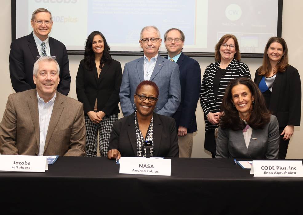 3 people sit behind a table covered with a black cloth. A group stands behind them for the Mentor-Protégé Agreement signing. 