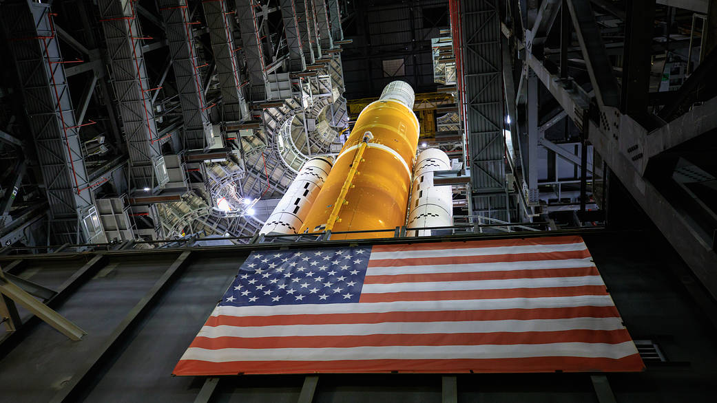Space Launch System rocket poised in the Vehicle Assembly Building with the American Flag on the Mobile Launcher in the foreground.