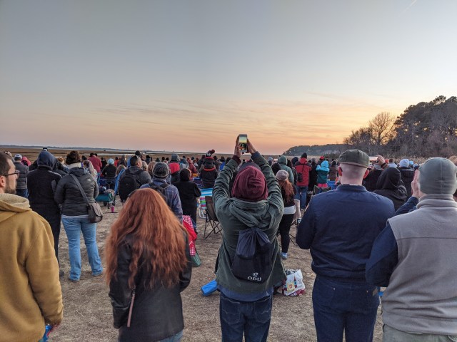A group of people stand facing away from the camera some holding cell phone cameras above their heads waiting to watch a rocket launch from Wallops Island
