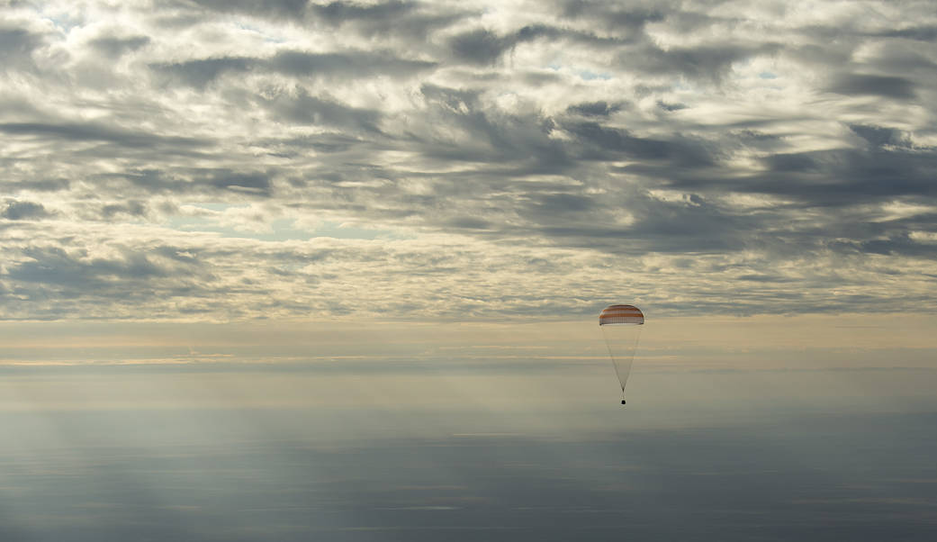 Soyuz capsule descends through clouds toward earth with parachute overhead