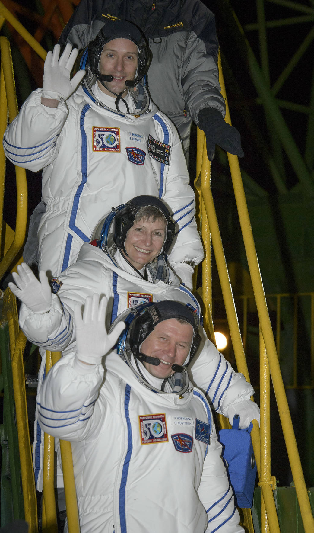 Three crew members in Sokol suits wave from ladder leading to Soyuz capsule