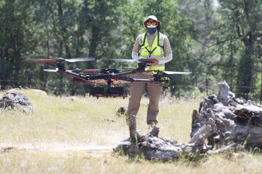 A drone flies in the foreground while a person wearing a hat, sunglasses and fluorescent yellow vest pilots the drone in the background, with a forest behind.