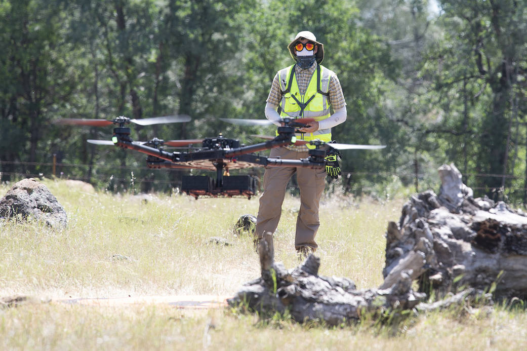 A drone flies in the foreground while a person wearing a hat, sunglasses, and yellow safety vest pilots the drone in the background, with a forest behind.