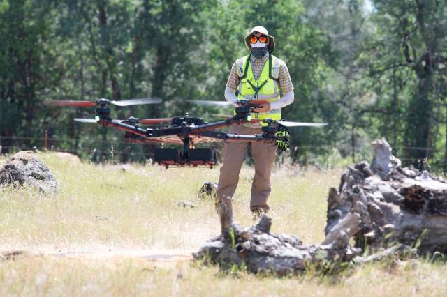 A drone flies in the foreground while a person wearing a hat, sunglasses, and yellow safety vest pilots the drone in the background, with a forest behind.