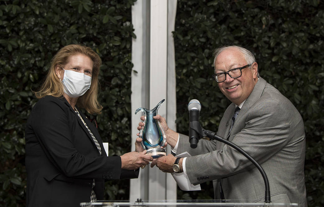 Woman wearing facemask, accepts an award from a man at podium with microphone.