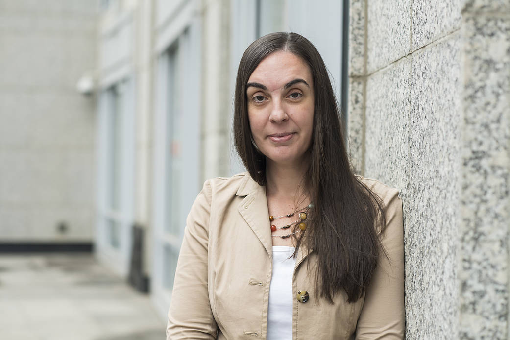 Jena Garrahy leans against the marble outside NASA headquarters wearing a beige jacket and white shirt, smiling at the camera with her mouth closed and hair down. 