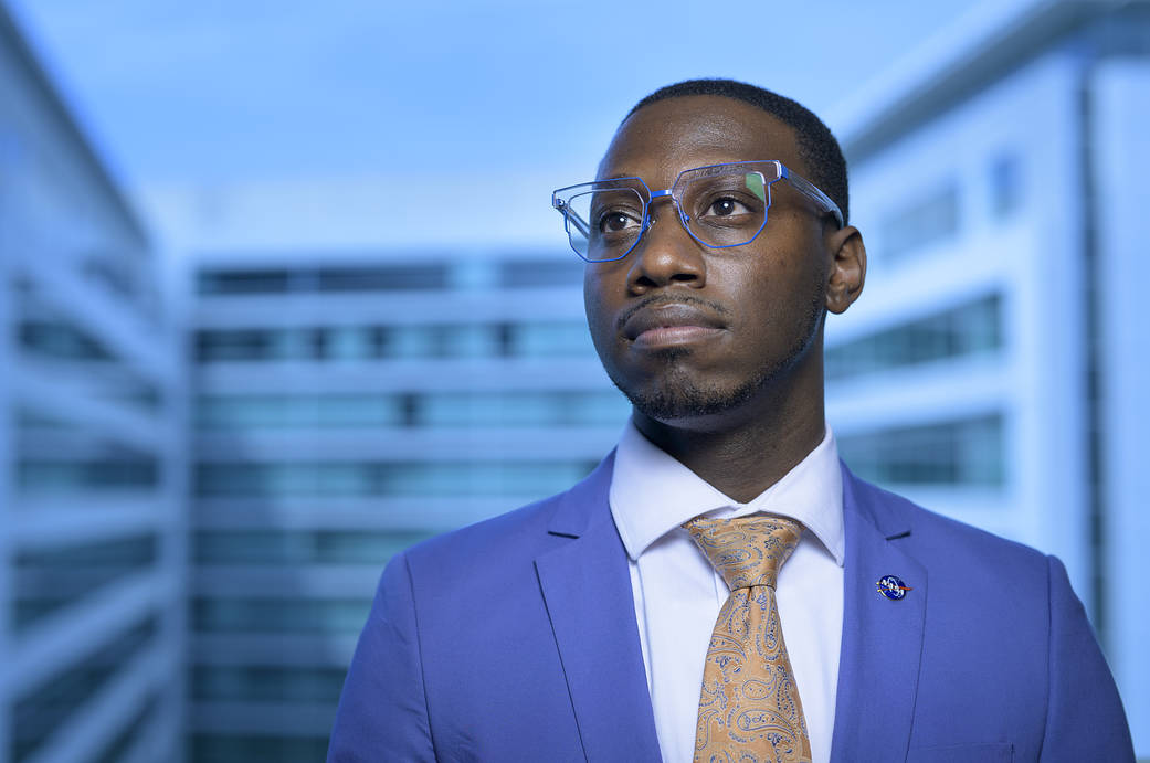Tyrone McCoy wears a crisp blue suit, light blue button up shirt, and orange and blue paisley tie, looking off into the distance to his right. There's a heroic feel to the captured moment, and the rims of his clear glasses reflect the blues in the scene. 