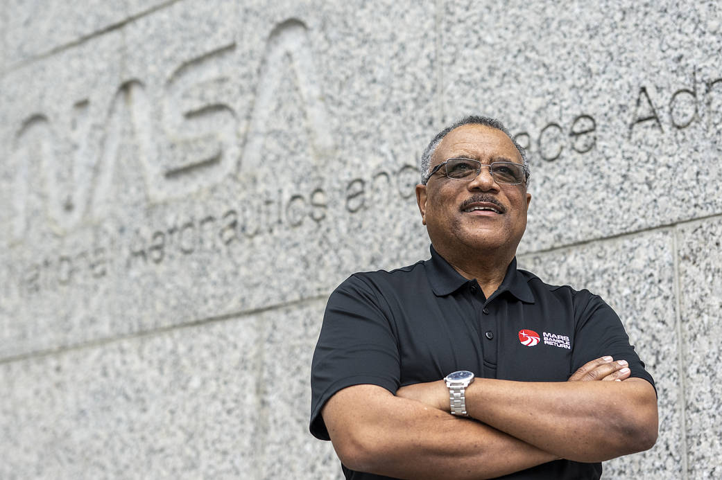 Dewayne looks off camera smiling with his arms crossed while wearing glasses, a silver watch, and a black collared shirt with the Mars Sample Return logo. He has a black mustache & salt & pepper short, curly hair. The NASA worm logo is in the background.
