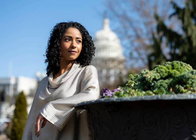Tahira Allen leans on a wall while gazing into the distance. She is wearing a white plaid sweater. A faint outline of the United States Capitol building can be seen in the background. 