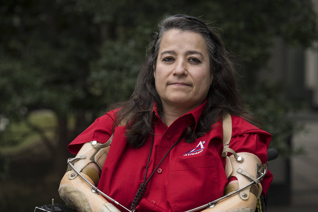 Dana smiles softly, wearing a red, short-sleeved collared shirt with the Artemis logo on it. She has long, dark brown hair and black eyeliner accentuating her dark brown eyes. She's seated in an electric chair, wearing her flesh-colored prosthetic arms.