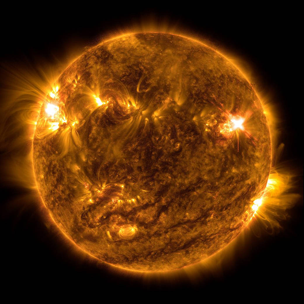 The Sun is gold with a mottled, swirling surface. Glowing particles burst out from the edge on the lower right and most of the upper left against the black background of space.