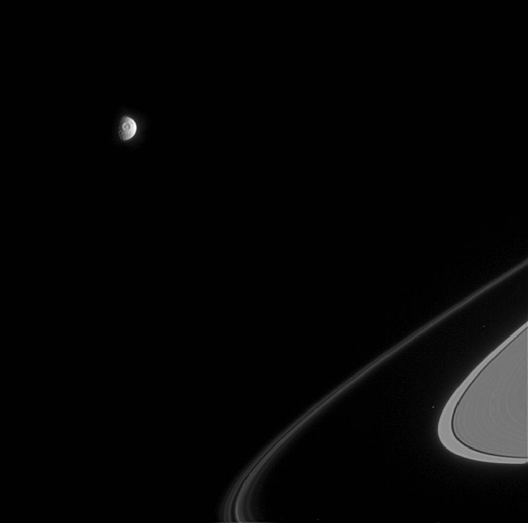 Saturn moon Mimas in distance with large crater visible and portion of Saturn rings at lower right