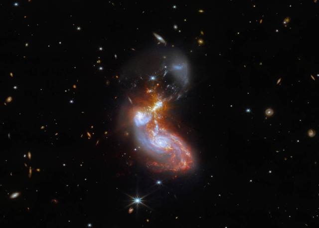 The two galaxies swirl into a single object in the center. Blue spiral arms stretch vertically. Gas spreads horizontally over that, mainly bright red with small gold spots. The core is bright and radiates eight large, golden diffraction spikes.