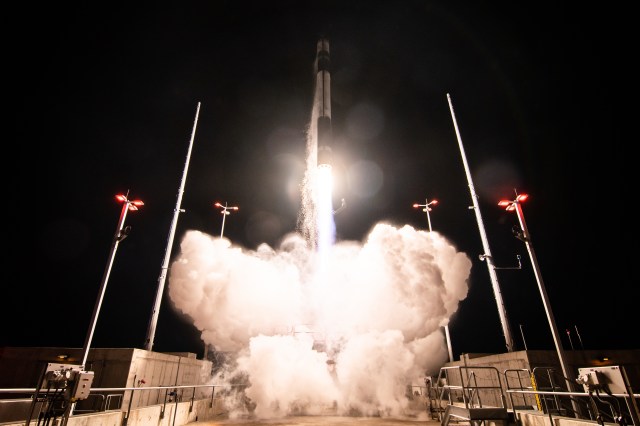 A nighttime photography of a black and white rocket just seconds launching off a launch pad, with a plume of bright white smoke just underneath.