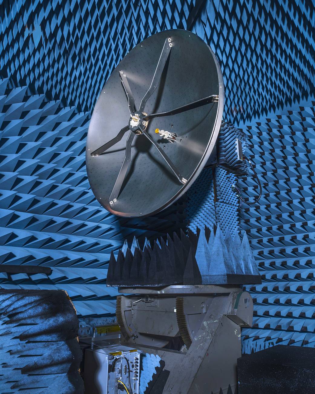 Roman’s high-gain antenna - a large, gray dish, about the height of a refrigerator, in a test chamber that is covered in blue spiked-shaped foam. A small circle is elevated in the middle of the antenna disk by six metal strips.