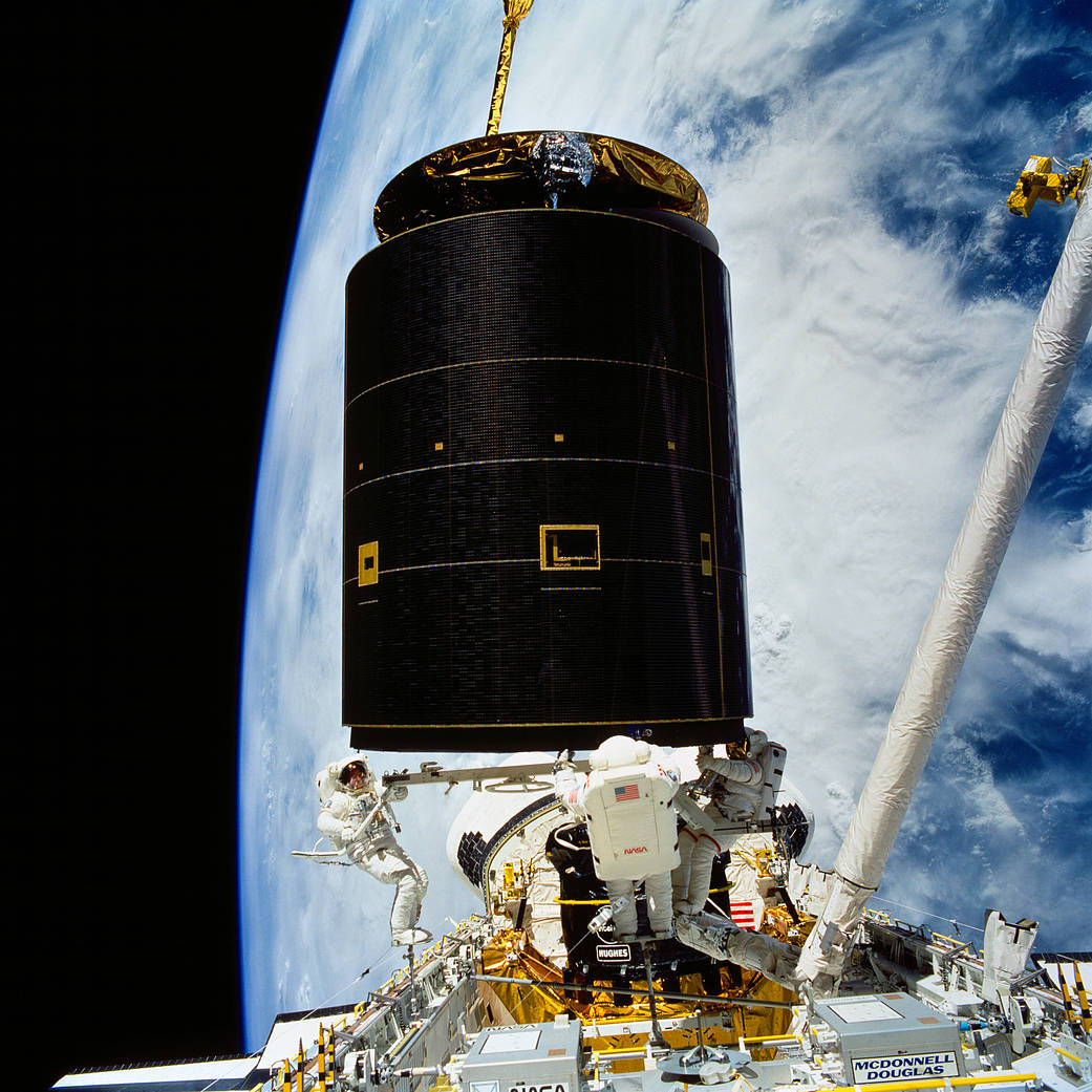 Three astronauts on a spacewalk maneuver a satellite into space shuttle cargo bay, with Earth visible in background