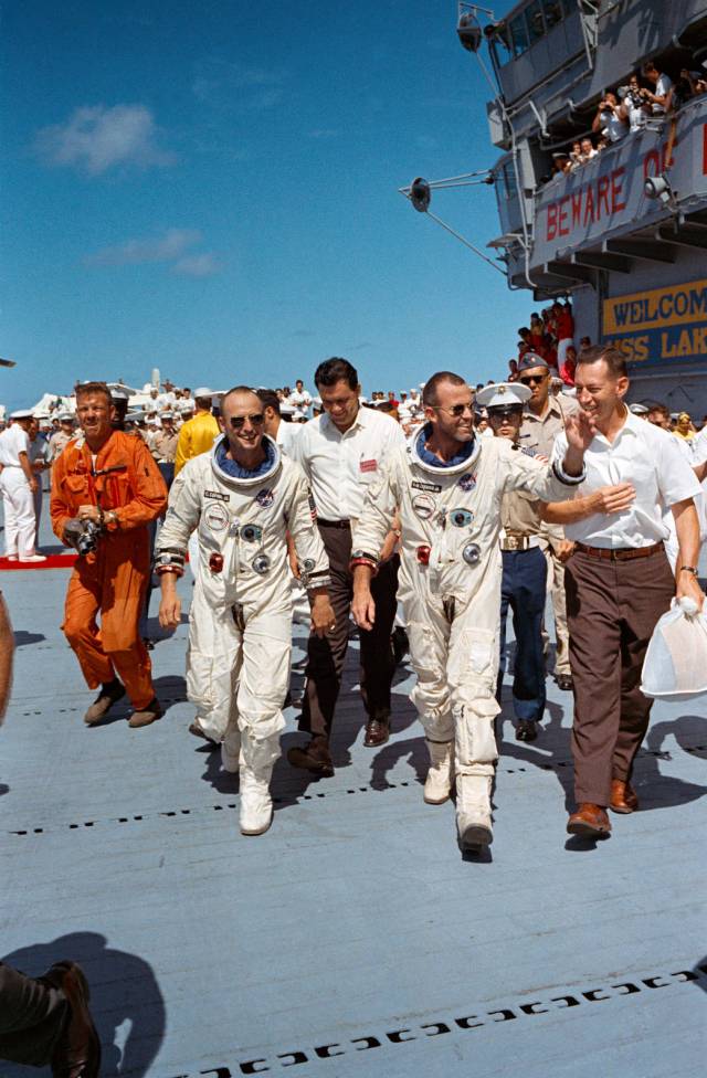 Two astronauts in flight suits walk across carrier deck, waving