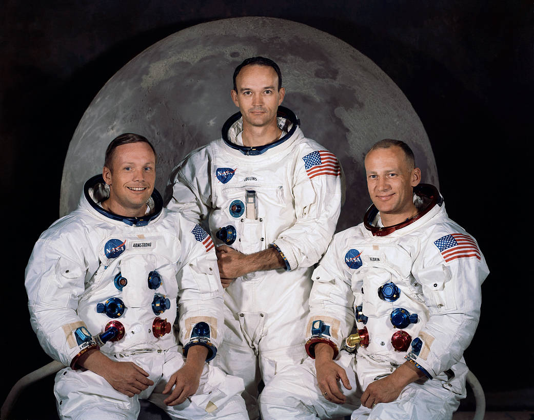 Three Apollo 11 astronauts in spacesuits without helmets in group portrait in front of Moon background