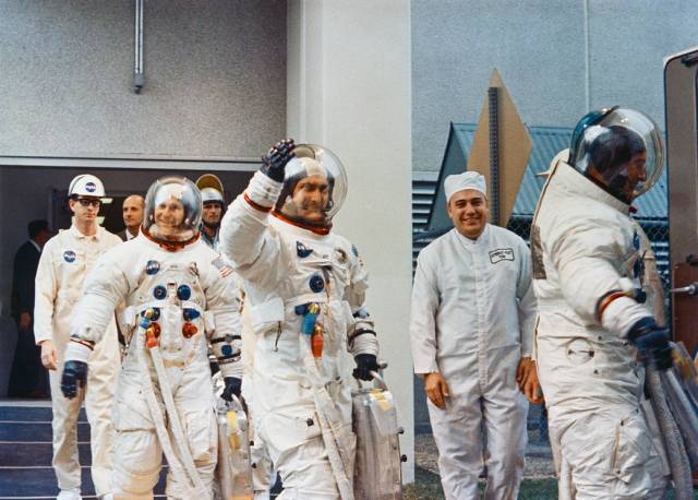 Three astronauts in spacesuits wave as they walk toward launchpad